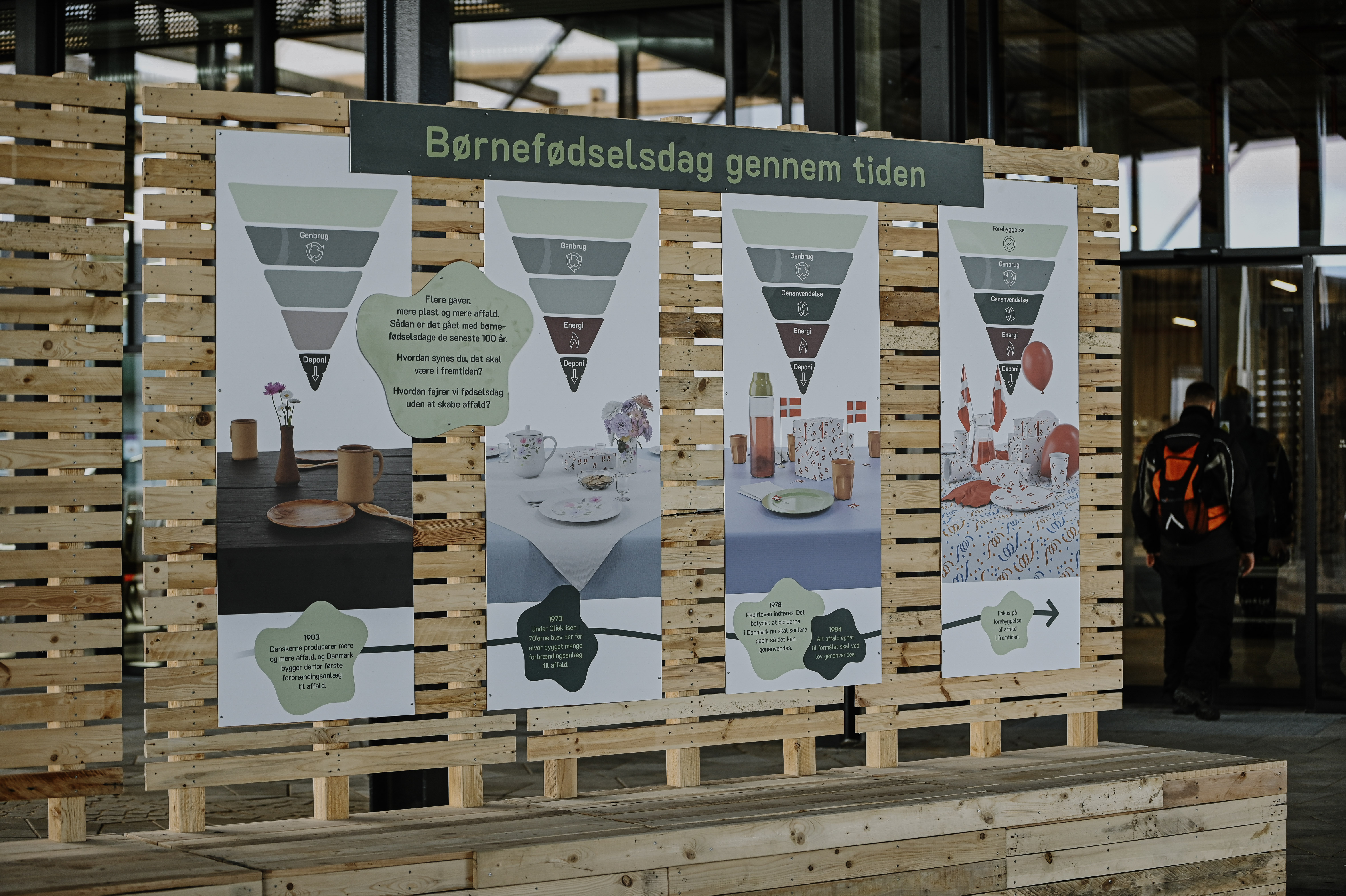 Udstilling på torvet i RessourceCenter Vejle (RCV) med temaet "Børnefødselsdag gennem tiden." Fire store informationsplakater er monteret på en træramme og viser udviklingen af fødselsdagsfejringer i forhold til bæredygtighed og affaldshåndtering. Hver plakat indeholder affaldshierarkier og billeder af forskellige bordopdækninger gennem tiden. I baggrunden ses indgangen til RCV, og en person med rygsæk er på vej ind.