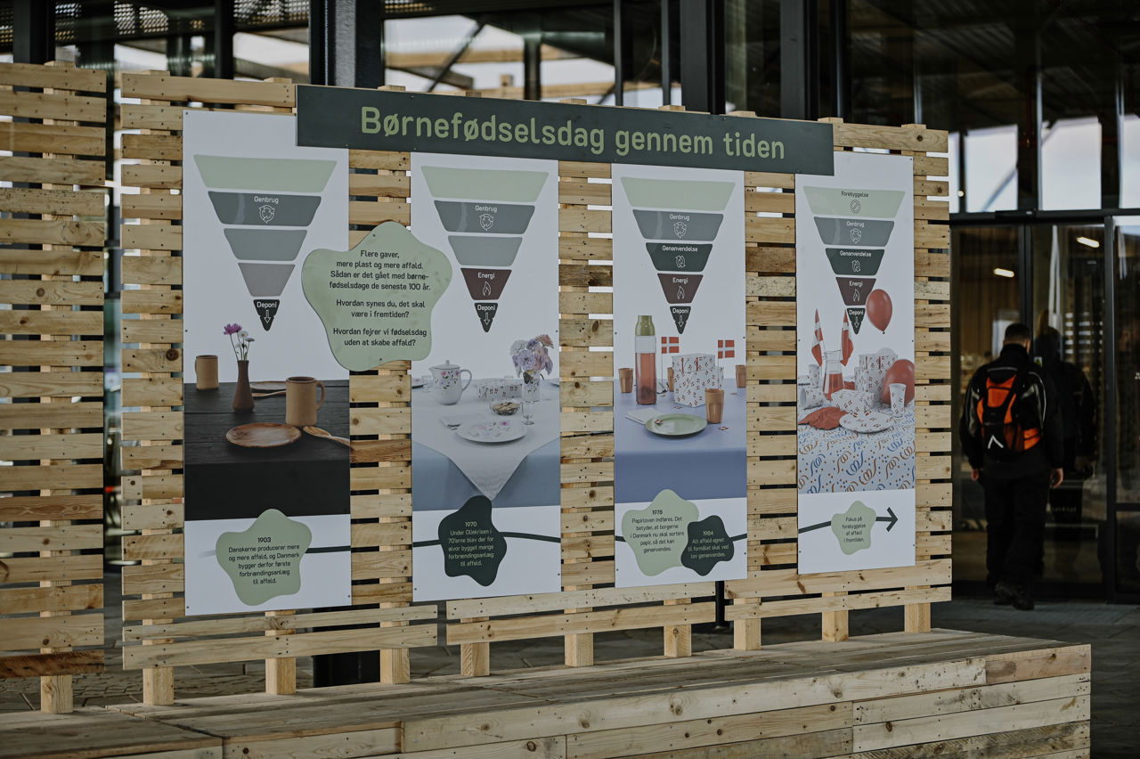 Udstilling på torvet i RessourceCenter Vejle (RCV) med temaet "Børnefødselsdag gennem tiden." Fire store informationsplakater er monteret på en træramme og viser udviklingen af fødselsdagsfejringer i forhold til bæredygtighed og affaldshåndtering. Hver plakat indeholder affaldshierarkier og billeder af forskellige bordopdækninger gennem tiden. I baggrunden ses indgangen til RCV, og en person med rygsæk er på vej ind.