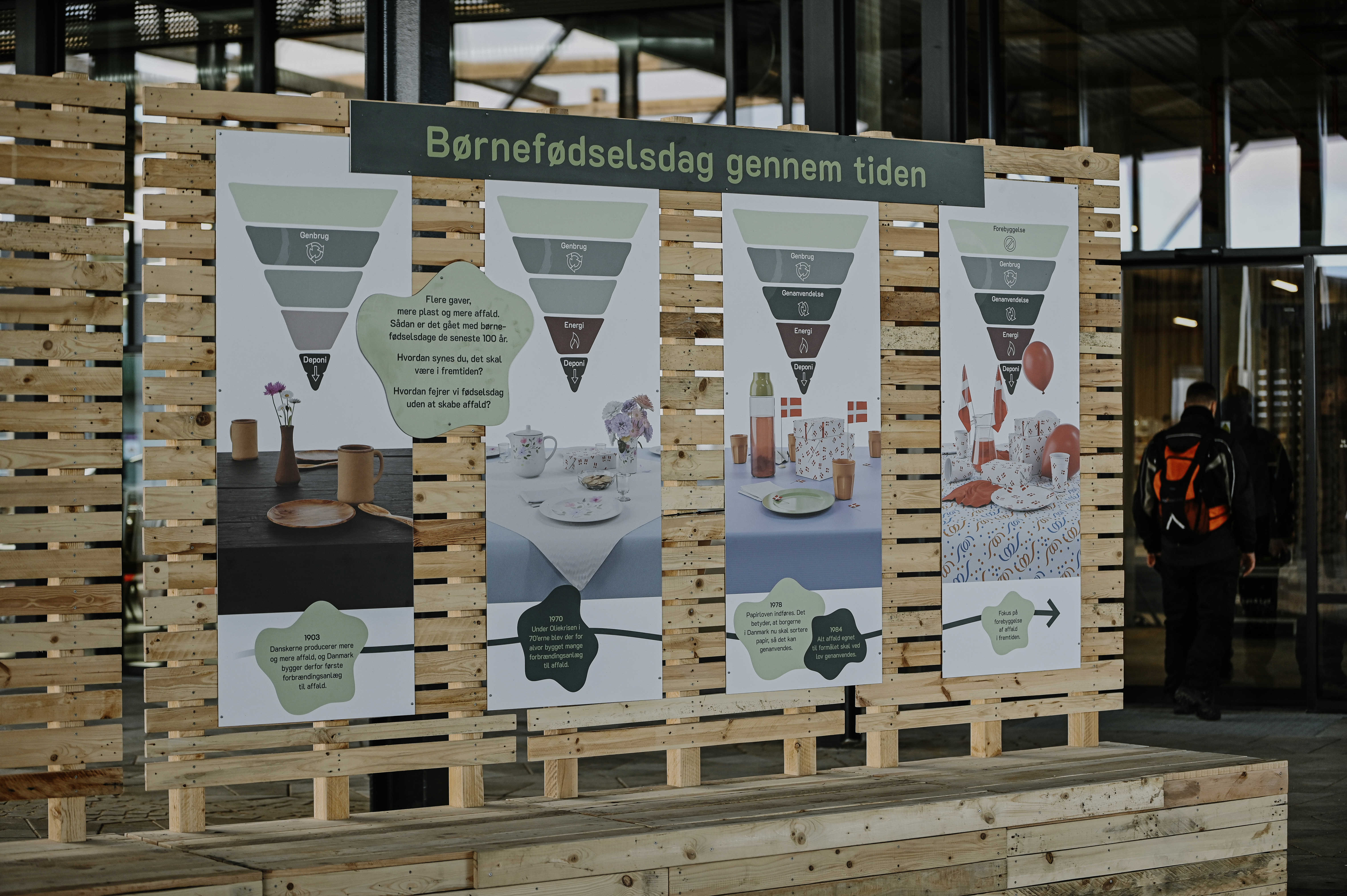 Udstilling på torvet i RessourceCenter Vejle (RCV) med temaet "Børnefødselsdag gennem tiden." Fire store informationsplakater er monteret på en træramme og viser udviklingen af fødselsdagsfejringer i forhold til bæredygtighed og affaldshåndtering. Hver plakat indeholder affaldshierarkier og billeder af forskellige bordopdækninger gennem tiden. I baggrunden ses indgangen til RCV, og en person med rygsæk er på vej ind.