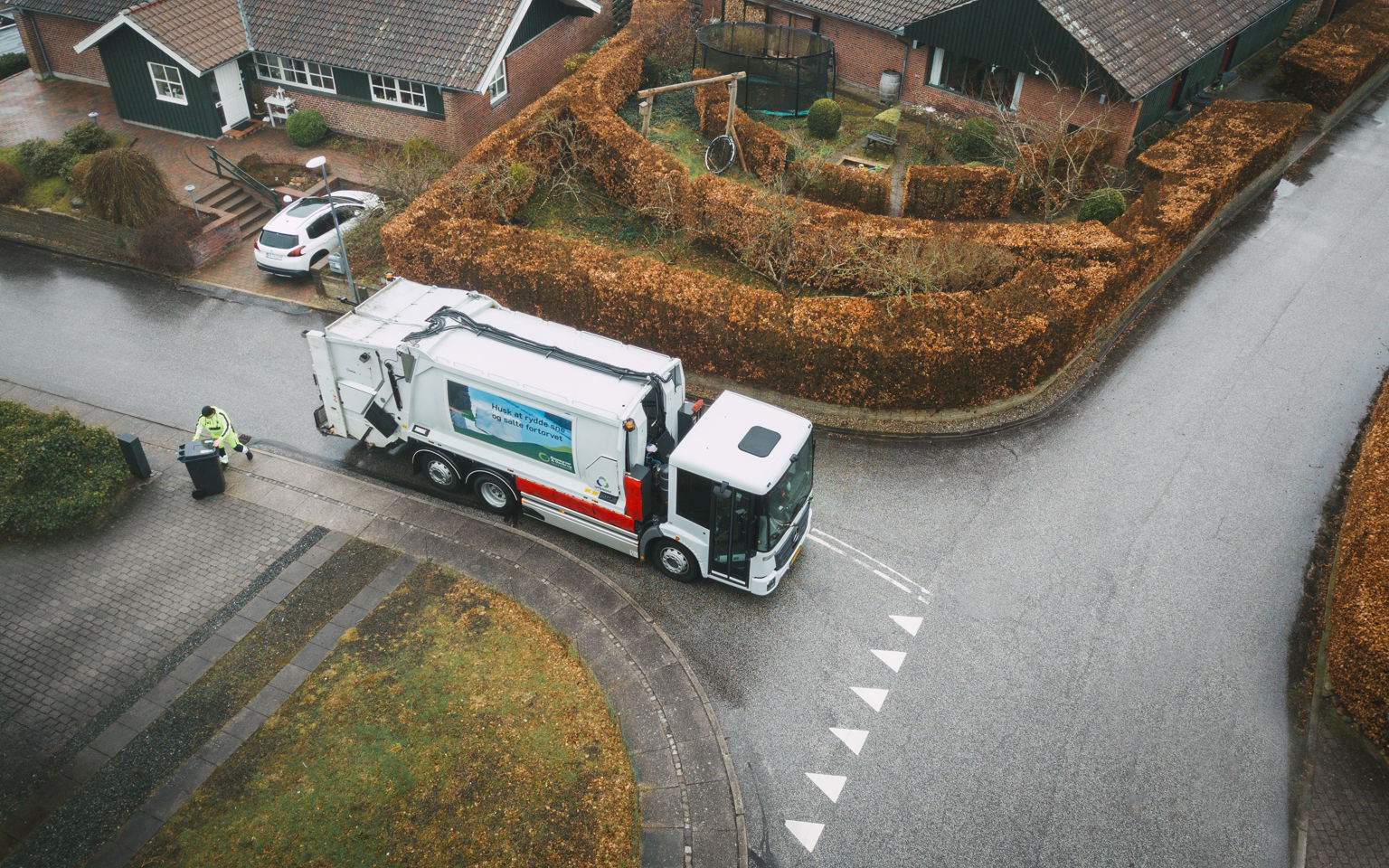 En skraldebil, der kører rundt i et boligområde med velplejede hække, mens en skraldemand tømmer en affaldsbeholder