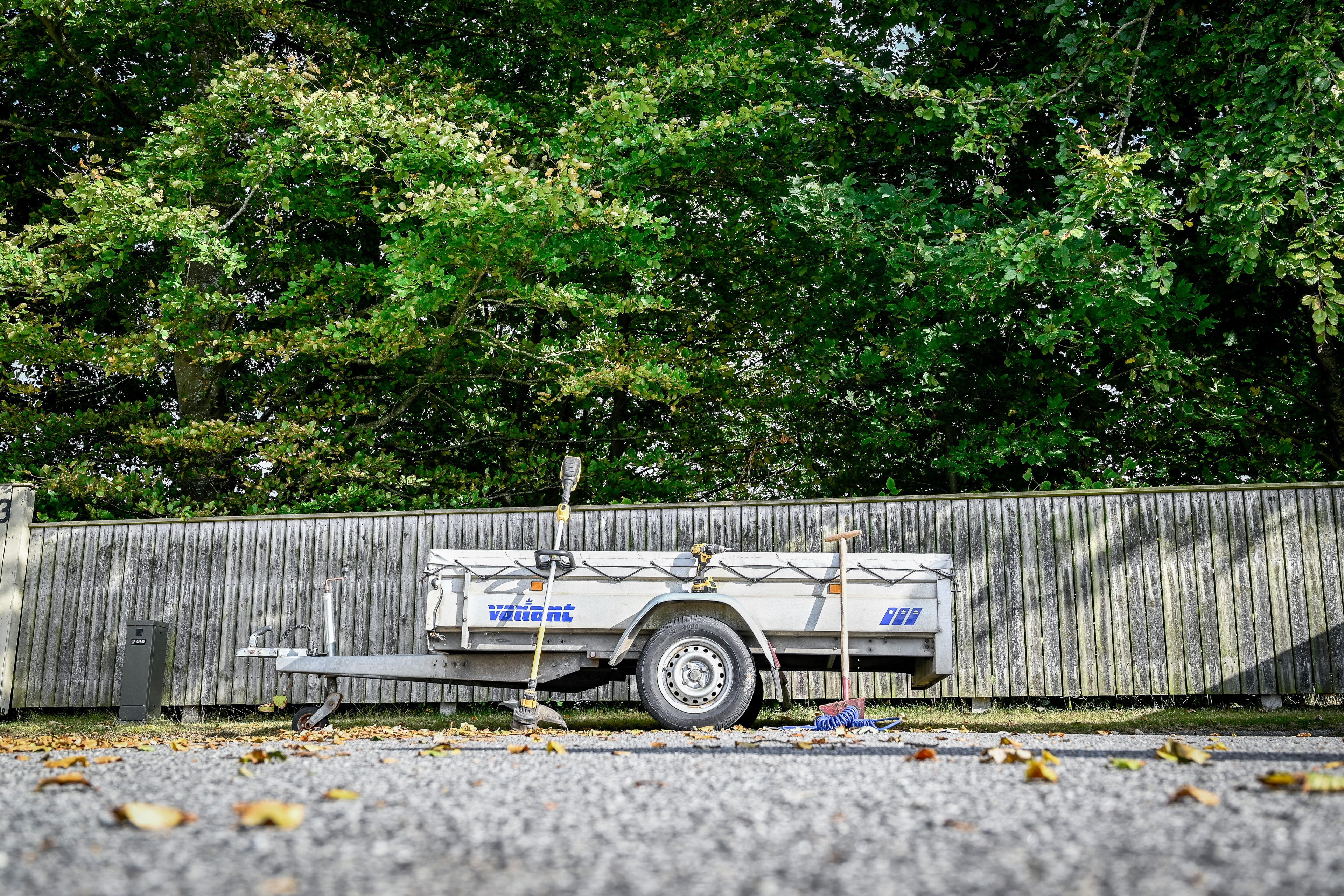Anhænger parkeret på villavej med forskellige haveredskaber og værktøj omkring. Bagved ses stakit og grønne trækroner.
