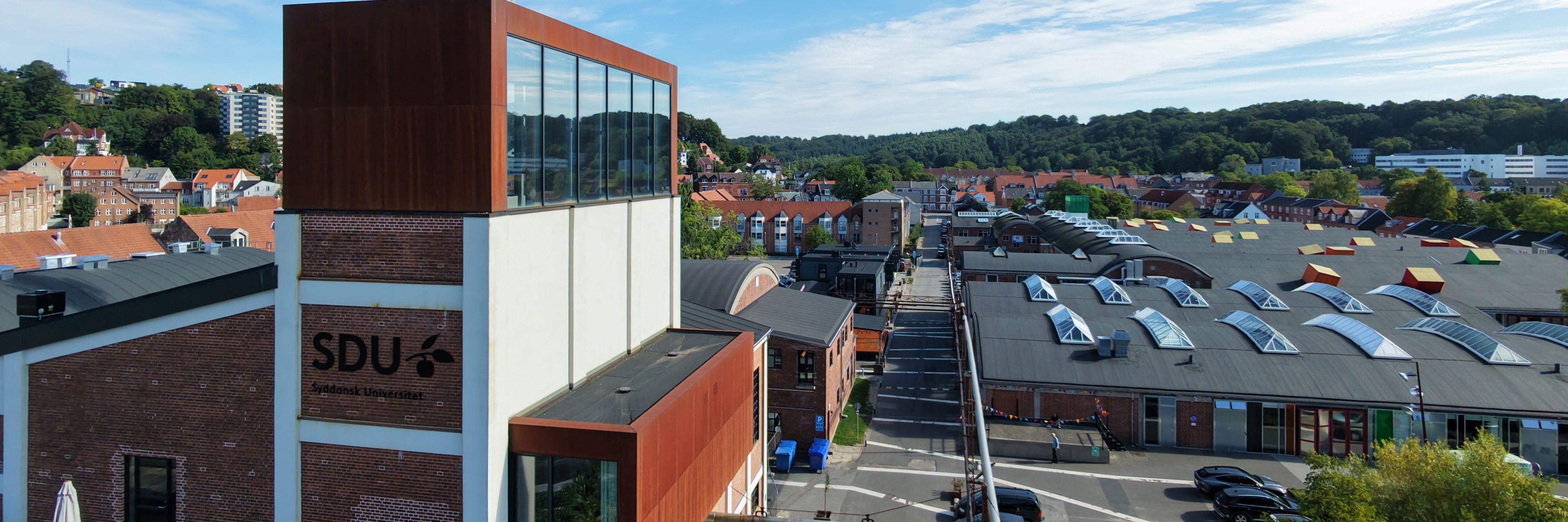 Dronebillede af Kedelbygningen med SDUs logo på siden. I baggrunden ses Spinderihallerne i Vejle.